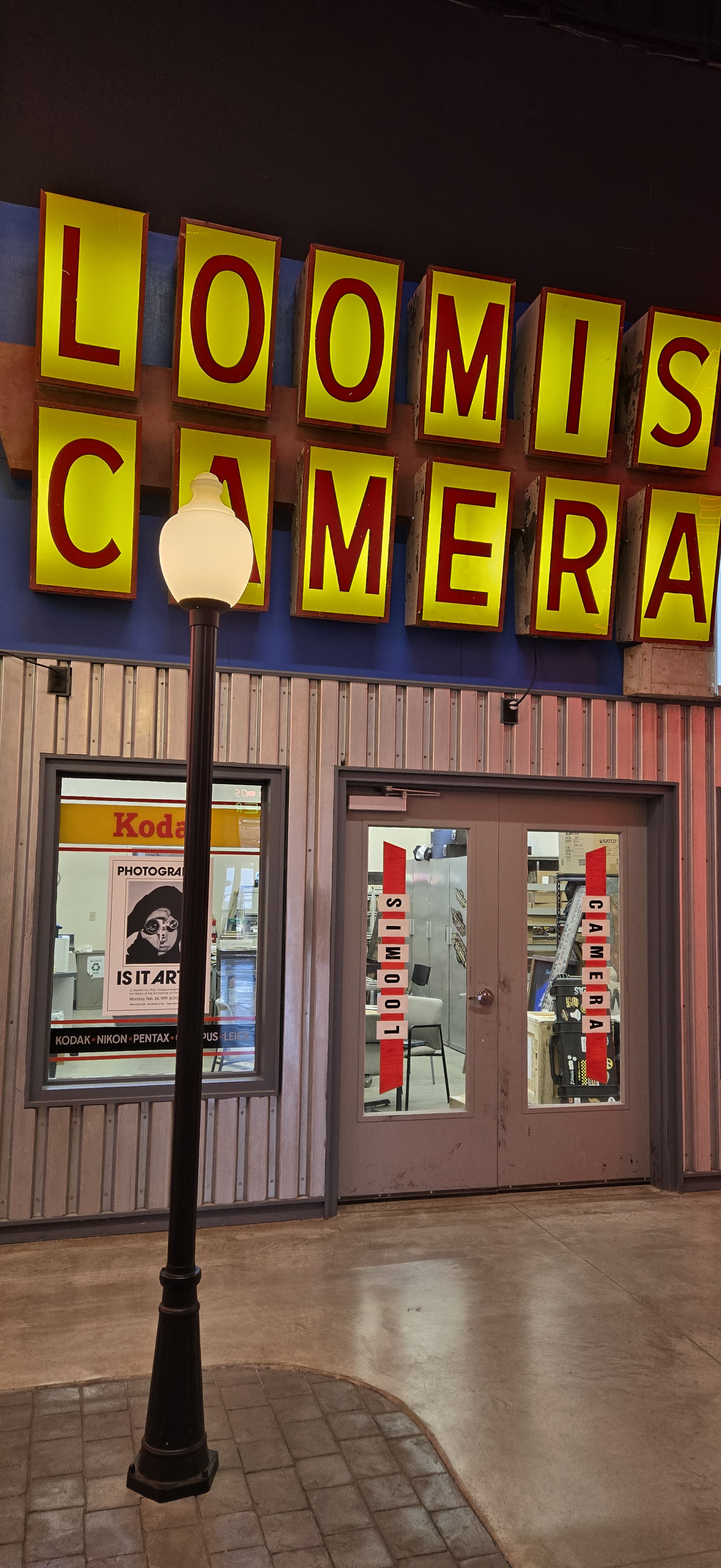 A vintage-inspired museum exhibit storefront labeled “Loomis Camera.” The large illuminated yellow and red sign hangs above a corrugated metal facade. Below it, glass double doors display vertical red “Loomis Camera” lettering. Through the doors, visitors can see into a workspace with shelves and equipment. To the left, a window displays a retro Kodak sign and a black-and-white photography poster reading “Is It Art?” A black streetlamp stands in front of the storefront on a cobblestone-style floor, evoking a nostalgic main street atmosphere.