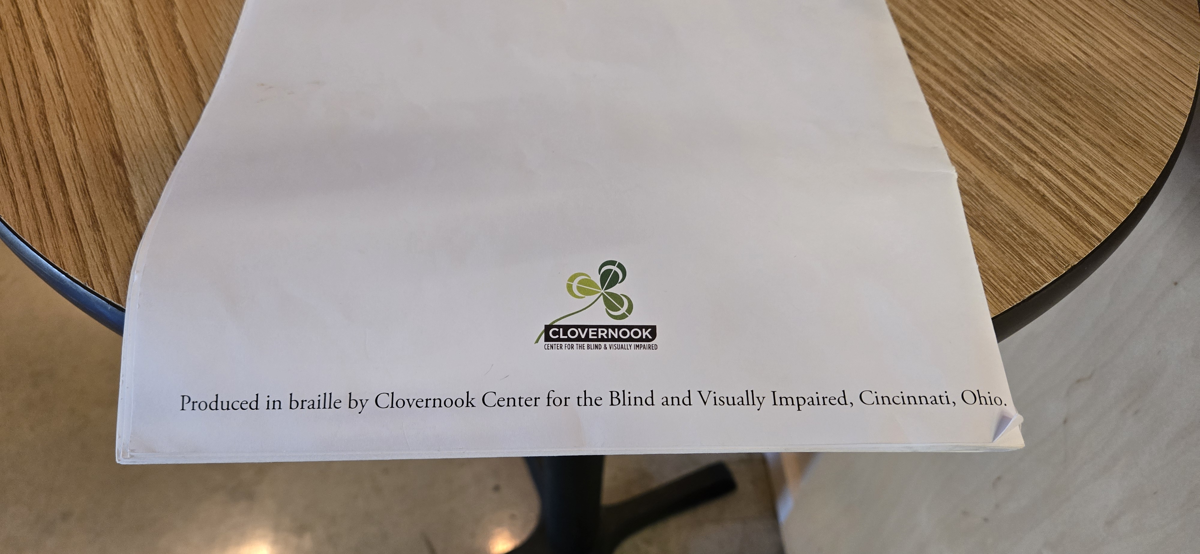 A white braille booklet resting on a wooden tabletop. The lower section shows the logo of Clovernook Center for the Blind and Visually Impaired, featuring a stylized green clover. Below the logo, printed text reads: “Produced in braille by Clovernook Center for the Blind and Visually Impaired, Cincinnati, Ohio.”
