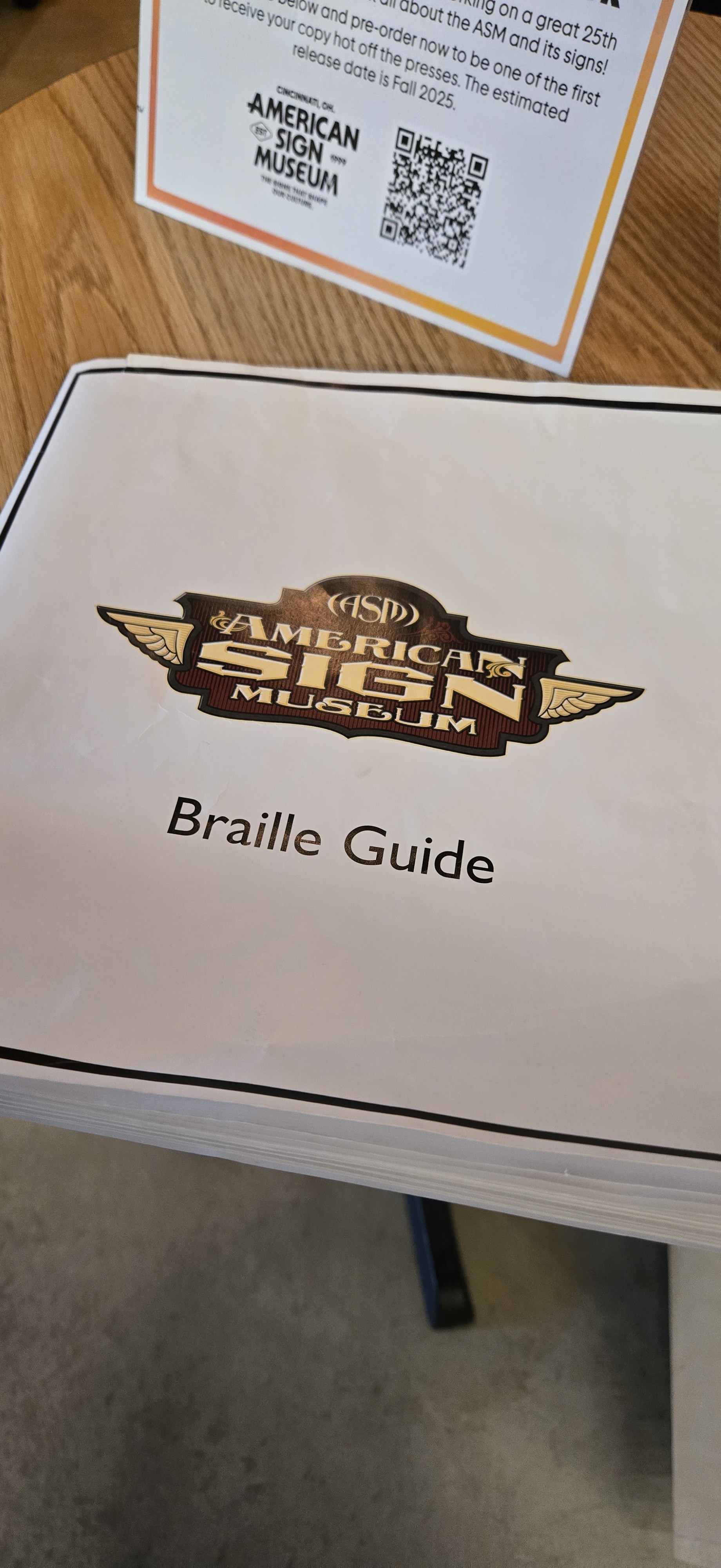 A white braille guide booklet from the American Sign Museum resting on a wooden table. The cover features the museum’s logo, styled with gold lettering and winged accents, reading “American Sign Museum.” Below the logo, printed text reads “Braille Guide.” In the background, a small informational sign promotes the museum’s upcoming 25th-anniversary book, featuring the museum’s logo, QR code, and preorder details.