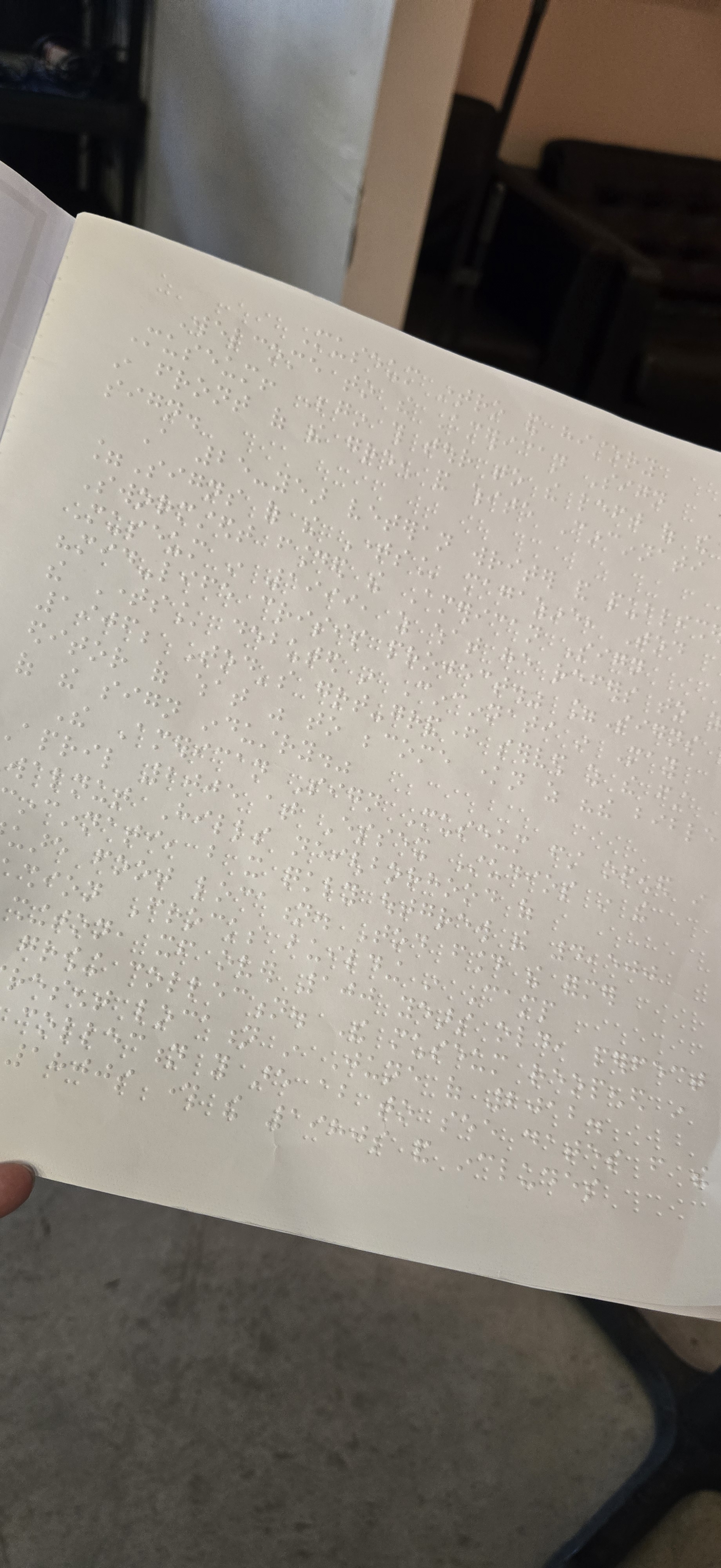 A close-up view of an open page from a braille guide at the American Sign Museum. The white page is covered with neatly arranged raised braille dots, allowing tactile reading. The background shows a dimly lit area with chairs and part of a table, emphasizing the focus on the accessible tactile text.