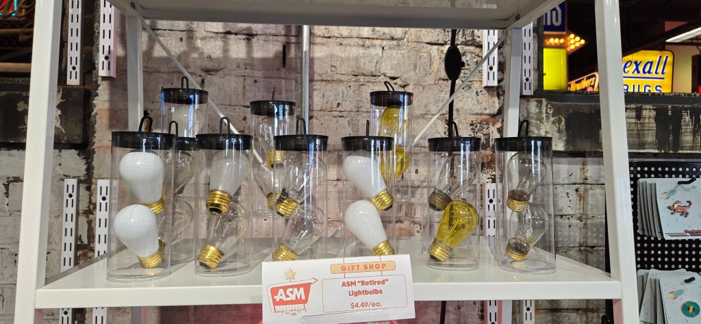 Display shelf in the American Sign Museum gift shop featuring clear plastic containers holding pairs of used lightbulbs labeled as “ASM ‘Retired’ Lightbulbs.” Some bulbs are white, others tinted yellow, with visible wear on the metal bases. A small sign in front lists the price as $4.49 each. Behind the display, a rustic brick wall and part of a vintage Rexall Drugs neon sign are visible in the background.