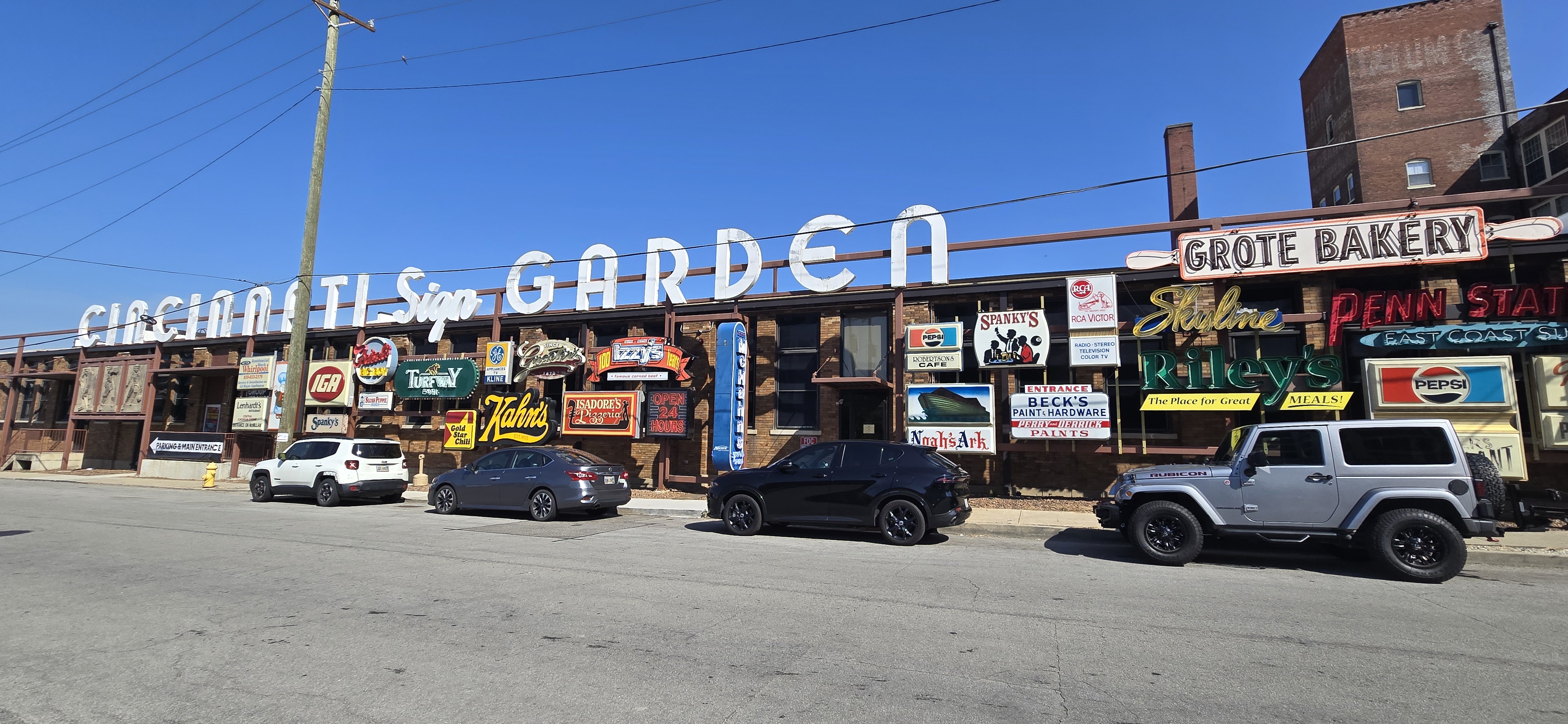 Exterior view of the American Sign Museum’s “Cincinnati Sign Garden,” a long brick building adorned with a large white rooftop sign reading “CINCINNATI Sign GARDEN.” The facade is covered with colorful vintage neon and painted signs from historic businesses, including Kahn’s, Gold Star Chili, Riley’s, IGA, Skyline, and Pepsi. Several parked cars line the street in front under a bright blue sky, emphasizing the lively display of preserved commercial signage.