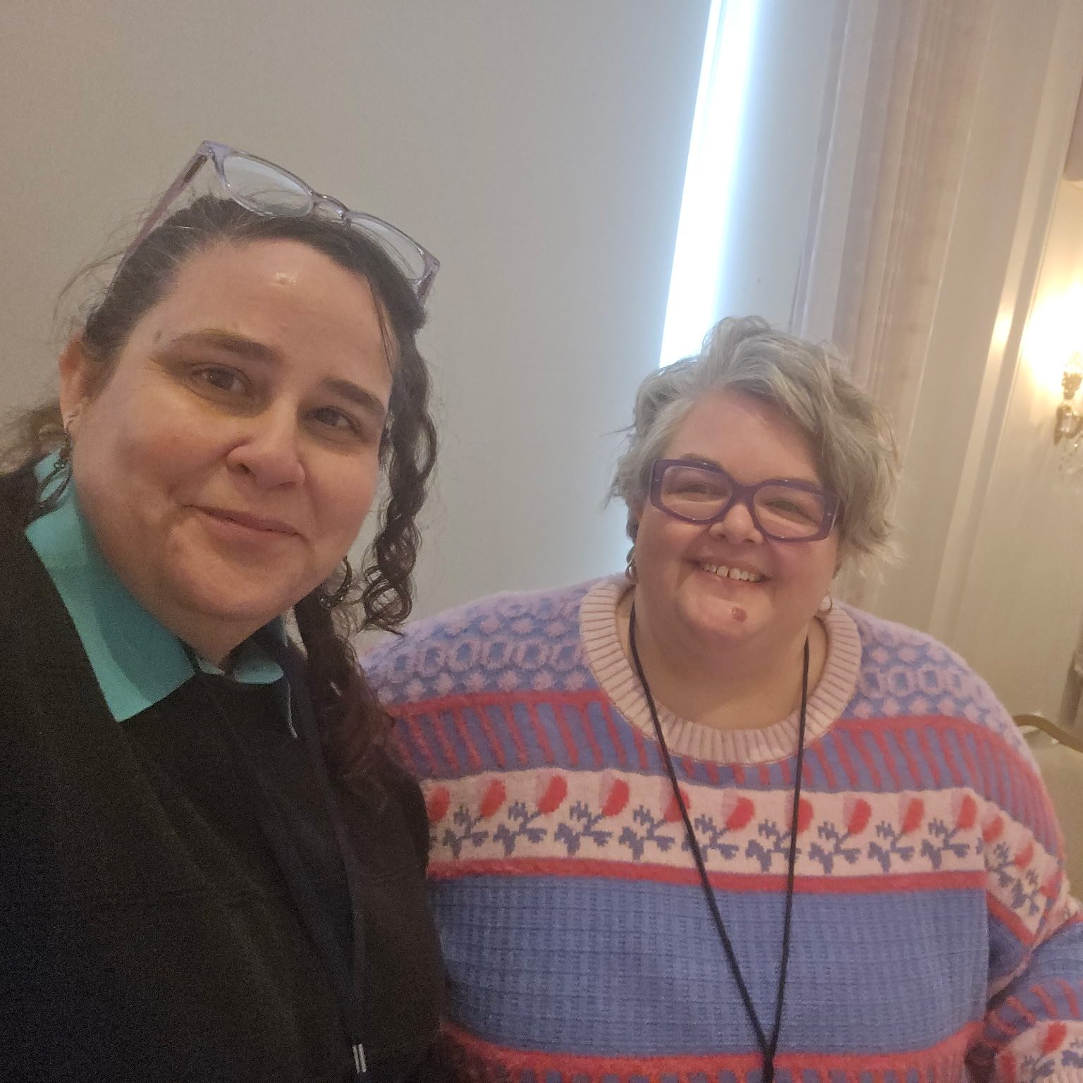 Two women smiling together at an event, with tables and elegant decor in the background.